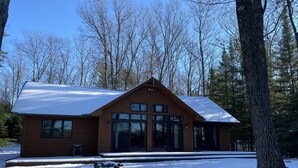 A wooden cabin with large windows and a snow-covered roof, surrounded by trees. - Sandy beach, quiet lake, great fishing & beautiful sunsets w/2 kayaks & gameroom (Cable)