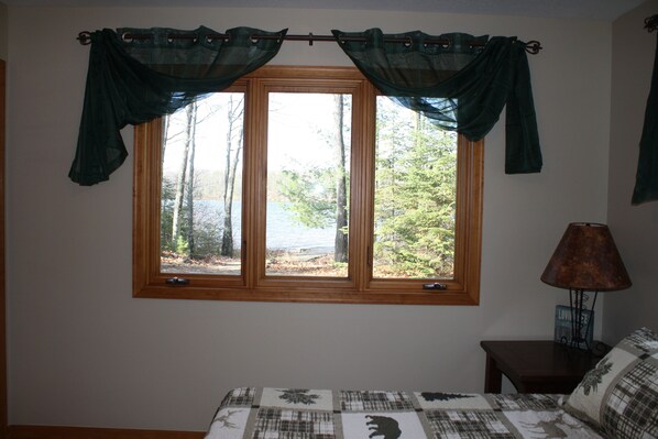 A bedroom with a large window, a bed with a patterned bedspread, and a nightstand with a lamp. - Sandy beach, quiet lake, great fishing & beautiful sunsets w/2 kayaks & gameroom (Cable)