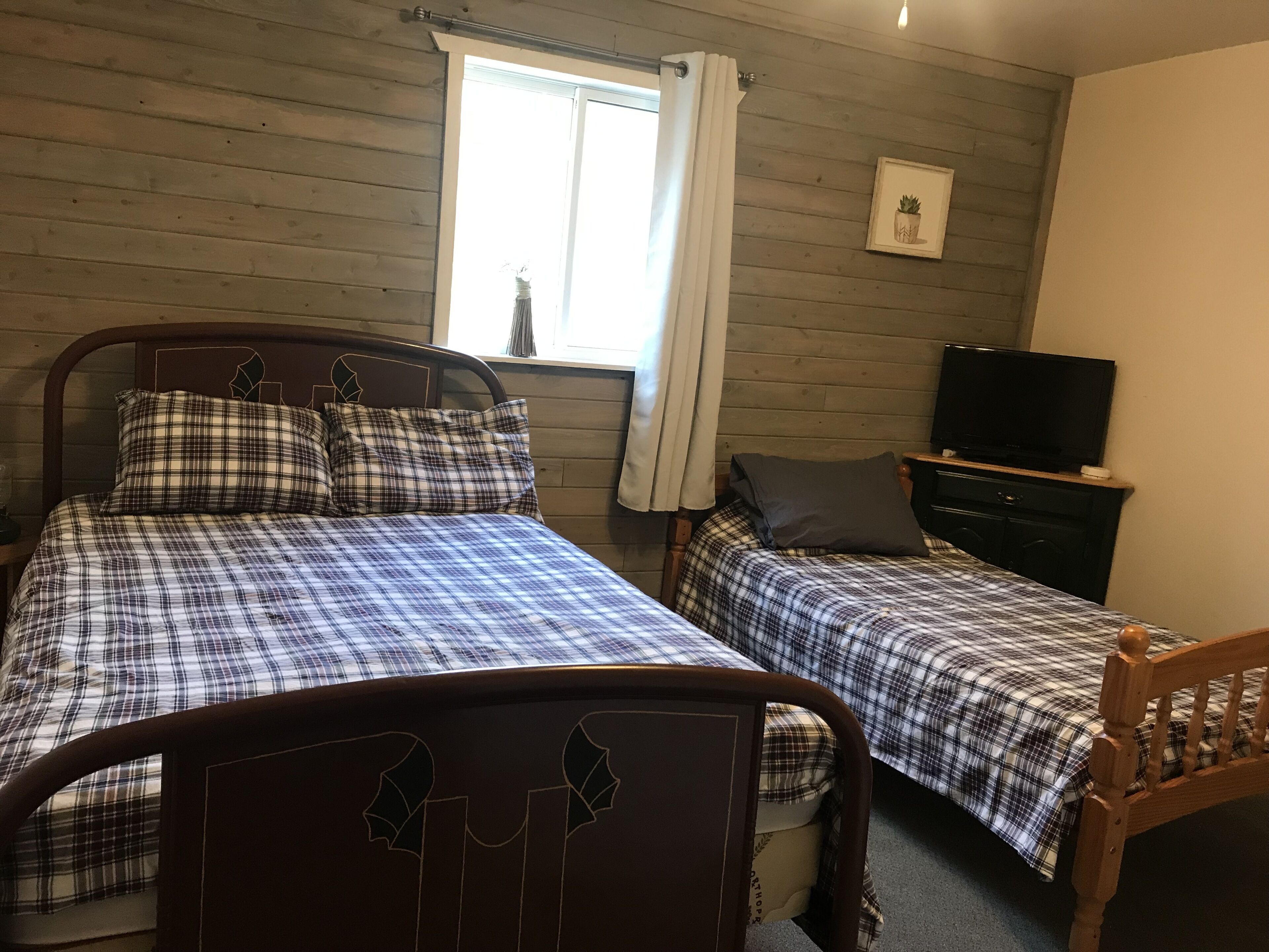A room with two beds, each with a wooden headboard and plaid bedding. There is a window with curtains, a television on a dresser, and a framed picture on the wall.