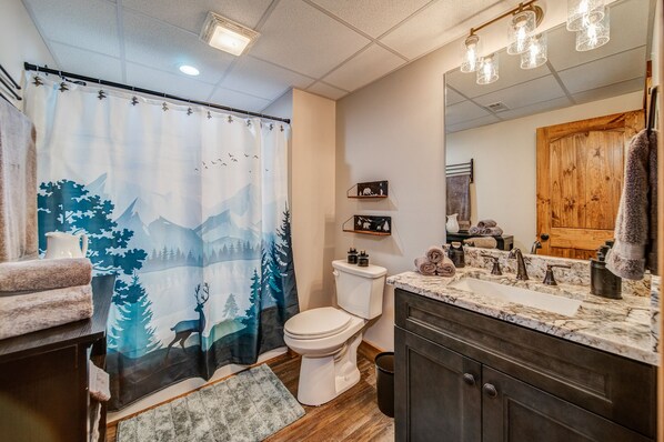 A bathroom with a shower curtain depicting a forest scene, a toilet, a sink with a marble countertop, and a wooden cabinet.