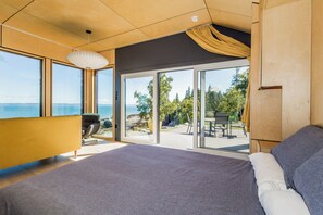 A modern bedroom with a large bed, a sofa, and a view of the outdoors through large windows. - Private Coastal Waterfall Escape, for Couples- Tides, Sauna, 55'  Ocean Falls (Canning)