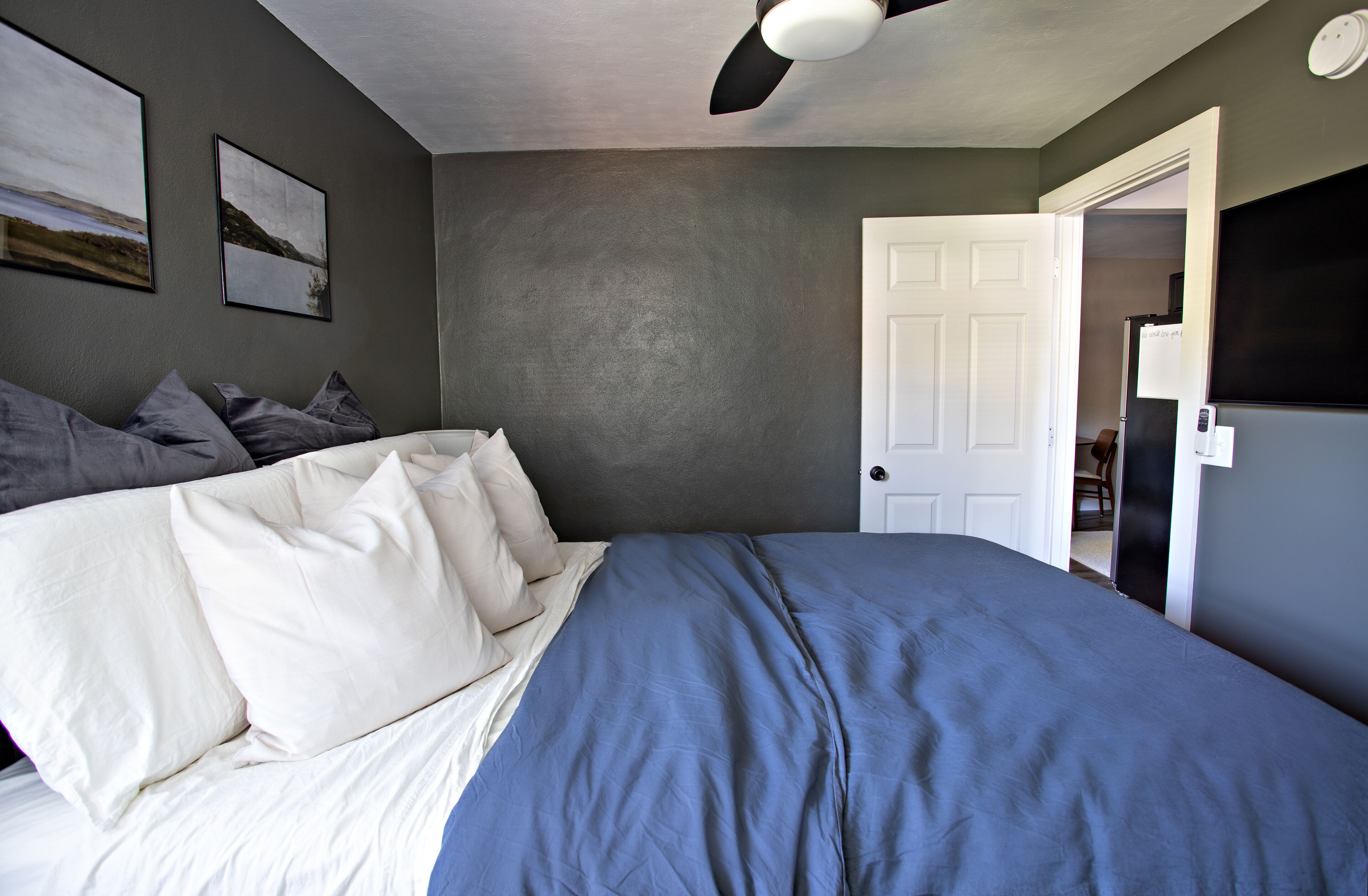 Een slaapkamer met een bed, kussens, een plafondventilator en een deur naar een andere kamer.