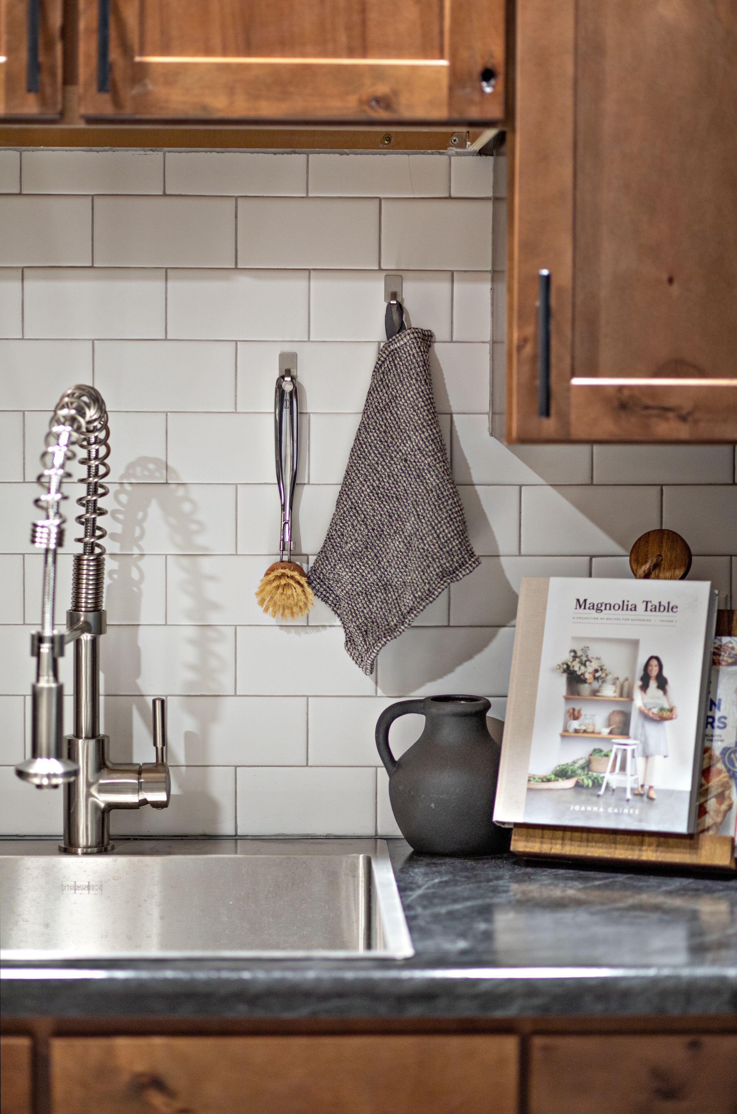 Una cocina con un lavamanos de acero inoxidable, una toalla colgante, una jarra negra y un libro de cocina titulado 'Magnolia Table' sobre la encimera.