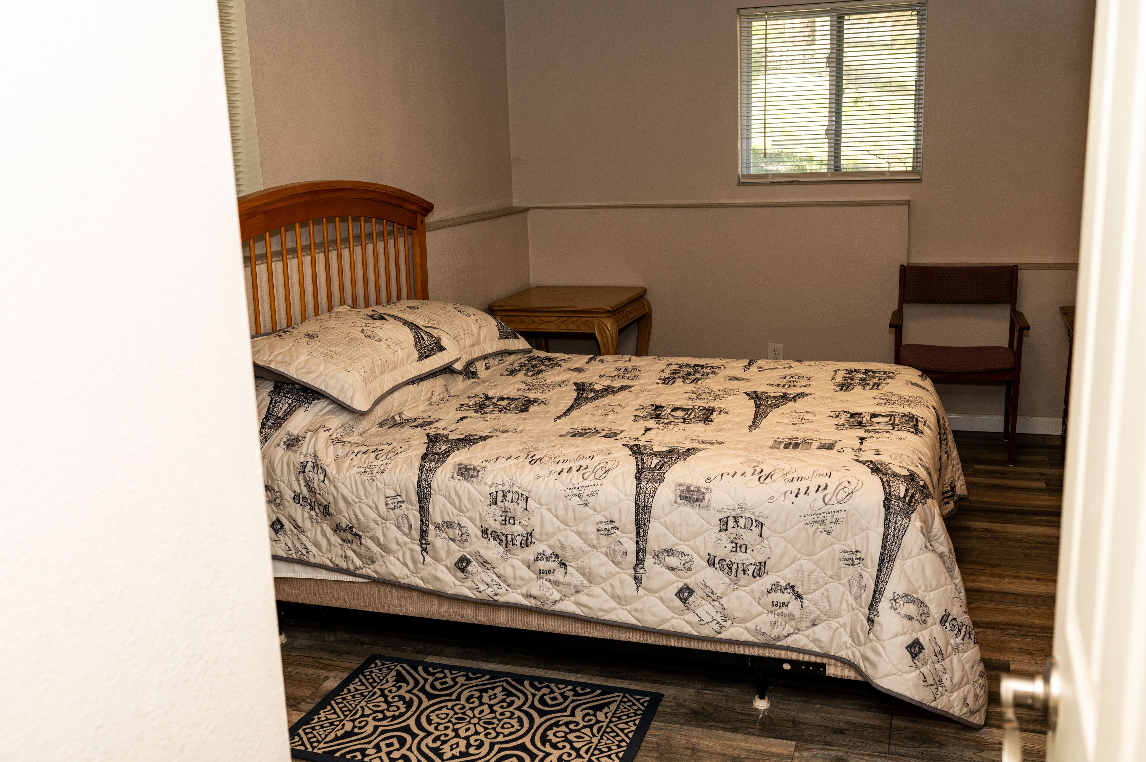 A bedroom with a bed, a wooden headboard, a small wooden table, a chair, a window, and a patterned rug.