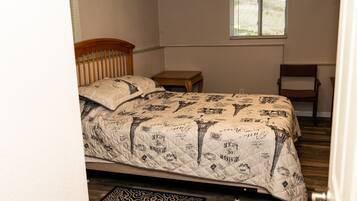A bedroom with a bed, a wooden headboard, a small wooden table, a chair, a window, and a patterned rug.
