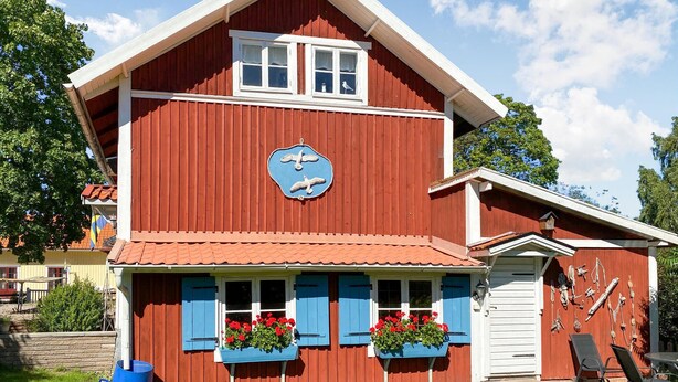 A red wooden building with a blue clock, two flower boxes, and a small porch area.