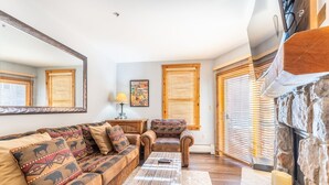 A cozy living room with a sofa, armchair, and coffee table, wooden blinds, and a stone fireplace. - 1BD IN THE HEART OF RIVER RUN VILLAGE - 2 MIN. WALK TO GONDOLA (Keystone)