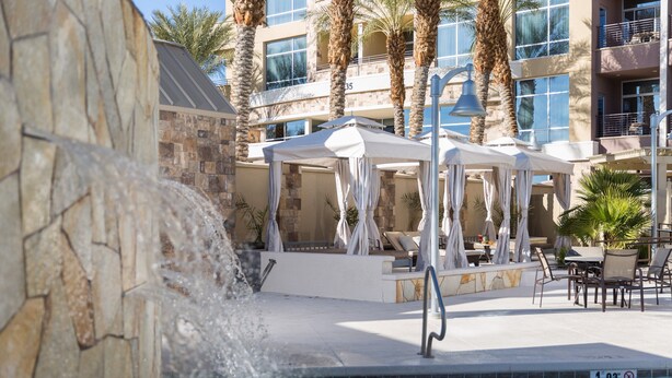 A hotel pool area with a stone wall, a water feature, lounge chairs, and umbrellas. - 1 Bedroom | Modern, Sophisticated Suite + Shuttles (Las Vegas)