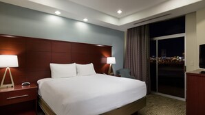 A hotel room with a large bed, two bedside lamps, a nightstand, a chair, a TV, and a view of the city at night. - 1 Bedroom | Modern, Sophisticated Suite + Shuttles (Las Vegas)