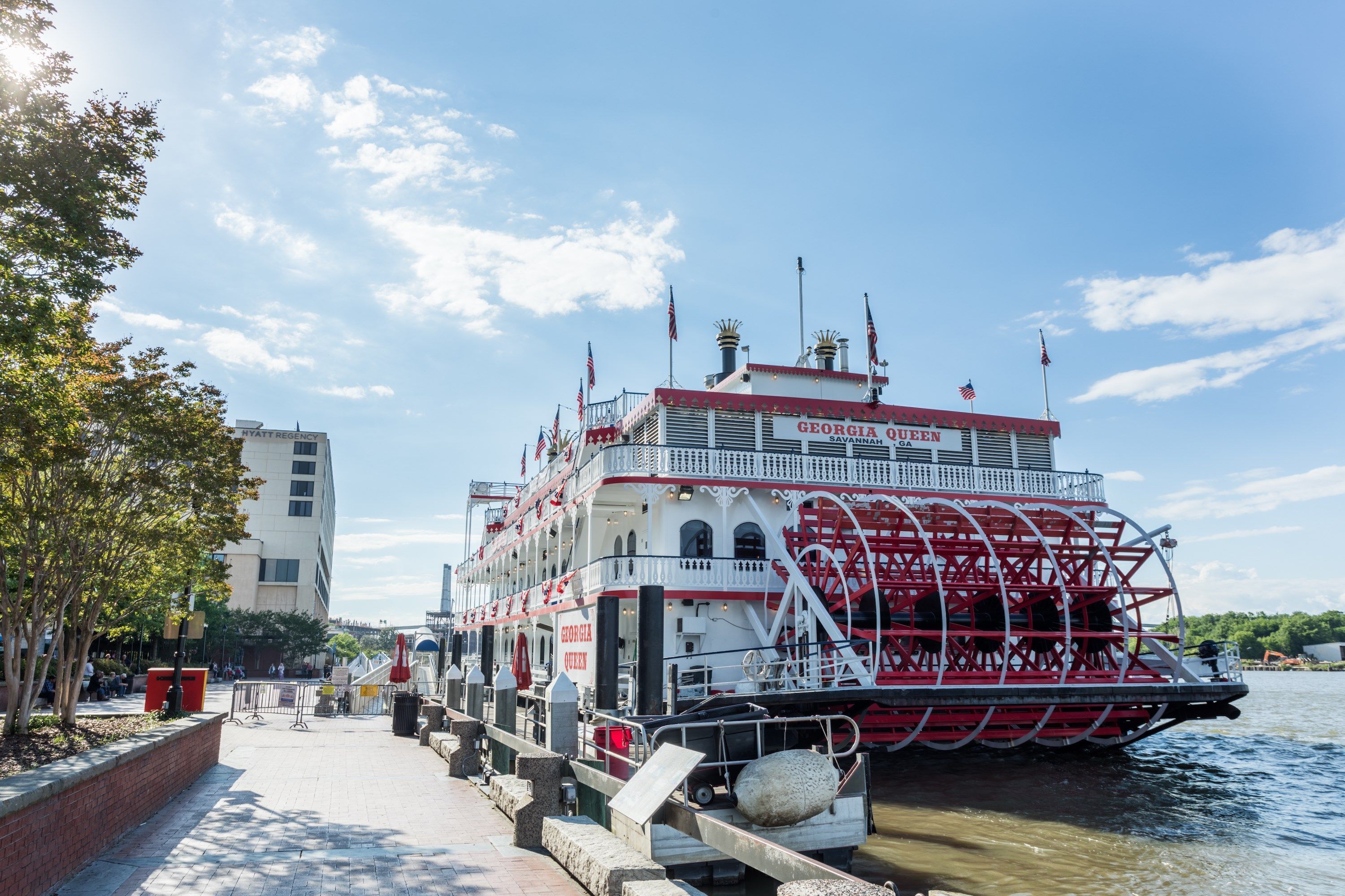 A large, red and white riverboat docked at a pier with a clear sky and trees in the background.