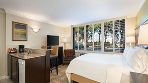 A hotel room with a bed, a desk with a chair, a television, a window with blinds, and a kitchen area.