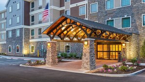A Staybridge Suites hotel with a prominent American flag, multiple windows, and a well-lit entrance. - 1 Bedroom | Breakfast Buffet. Pool & Hot Tub (Canton)