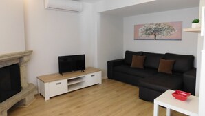 Ein modernes Wohnzimmer mit Kamin, einem Flachbildfernseher auf einem Holzständer, einer schwarzen Couch mit braunen Kissen und einem kleinen weißen Tisch mit einem roten Tablett.