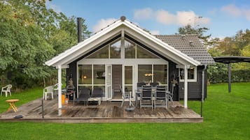 Une maison moderne avec une grande façade vitrée, une terrasse en bois et du mobilier d’extérieur.