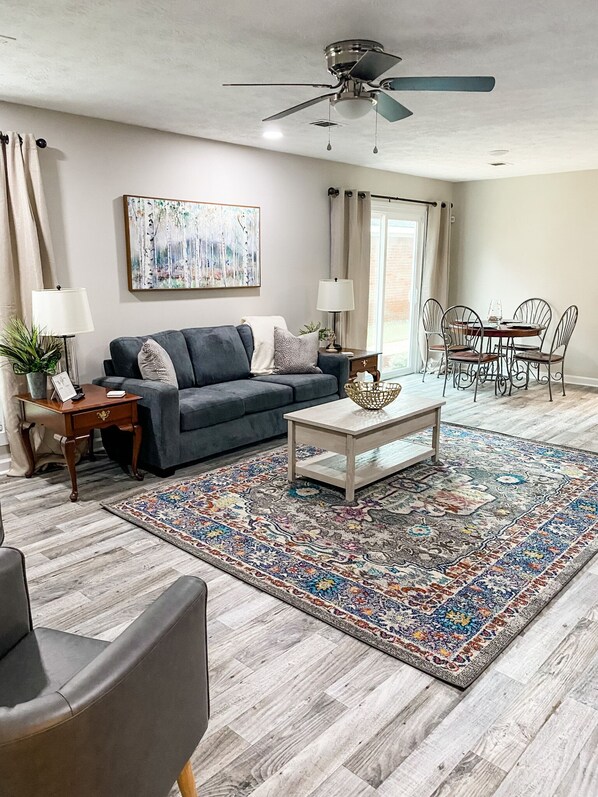 A living room with a grey sofa, a coffee table, a patterned rug, a dining area with metal chairs, and a ceiling fan.