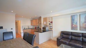 A modern kitchen with wooden cabinets, a dining area with a glass table, and a living room with a leather sofa.