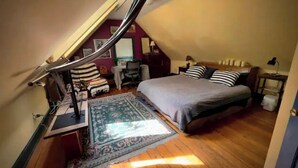 A cozy attic bedroom with a sloped ceiling, a bed with striped bedding, a desk with a lamp, and a patterned rug.
