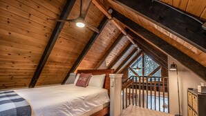 A bedroom with a sloped ceiling, a bed with a white comforter, a red pillow, and a view of the outdoors through a window.