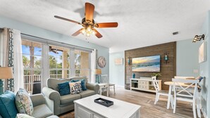 A spacious living room with a ceiling fan, a large window with a view, a dining area with white furniture, and a TV mounted on the wall. - First Floor • Walk to Beach • Sleeps 6 (Panama City Beach)