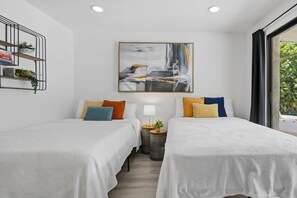 Quarto de hotel moderno com duas camas, um quadro na parede, uma mesinha com uma luminária e uma janela com vista para áreas verdes.