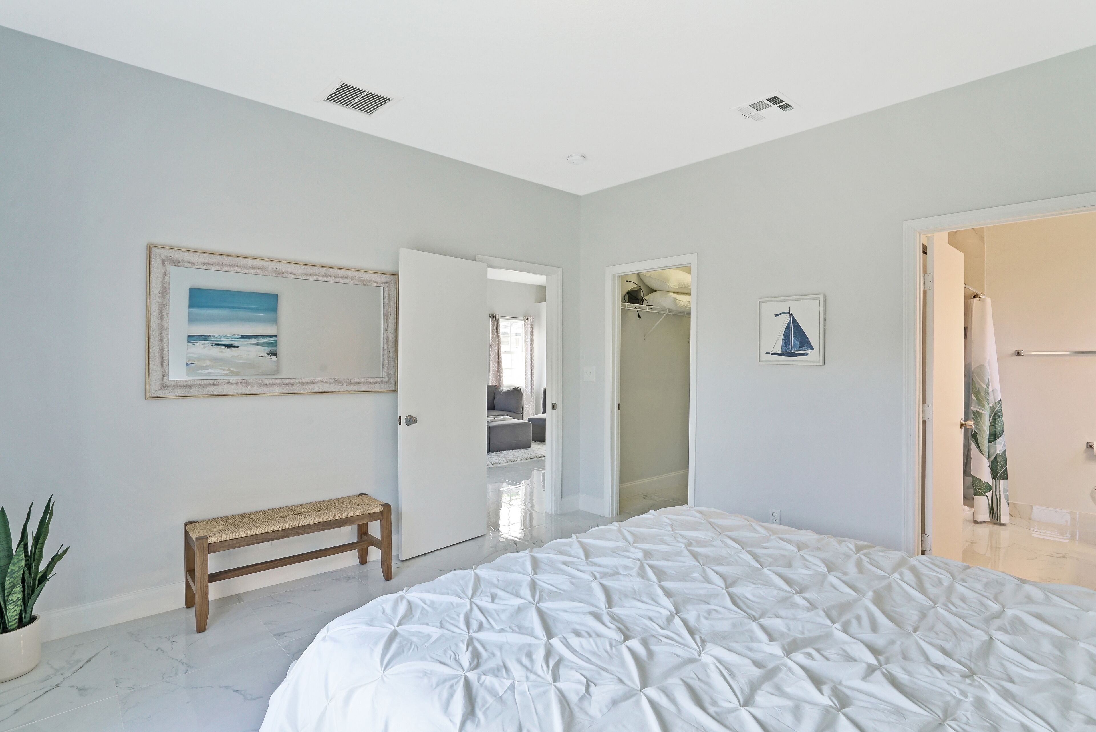 A bedroom with a large bed, a bench, a potted plant, and a door leading to another room.