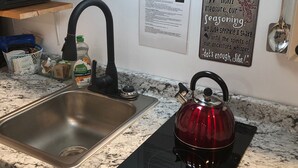 A kitchen with a stainless steel sink, a red kettle on an induction cooktop, and a sign on the wall. - Studio B. Katie and Steve Riley Apartment (Breaux Bridge)