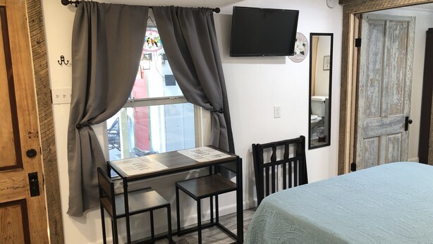 A room with a bed, a table with chairs, a TV on the wall, and a window with curtains. - Studio B. Katie and Steve Riley Apartment (Breaux Bridge)