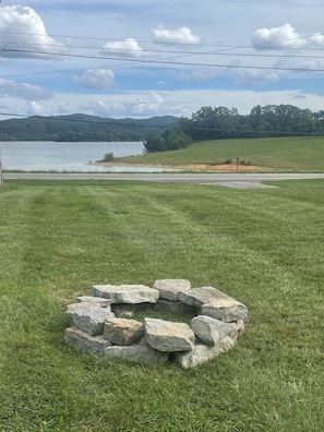 A grassy area with a stone fire pit in the foreground, a road, and a lake with trees in the background.