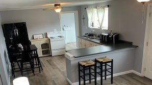 A modern living room with a grey sofa, a coffee table, and a patterned throw pillow. A kitchen with a white stove, sink, and bar stools is visible in the background.