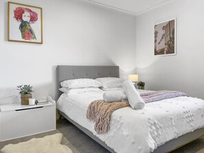 Une chambre à coucher avec une tête de lit recouverte d’un tissu gris, du linge de lit blanc, une lampe de chevet, une petite plante et un tableau encadré au mur.