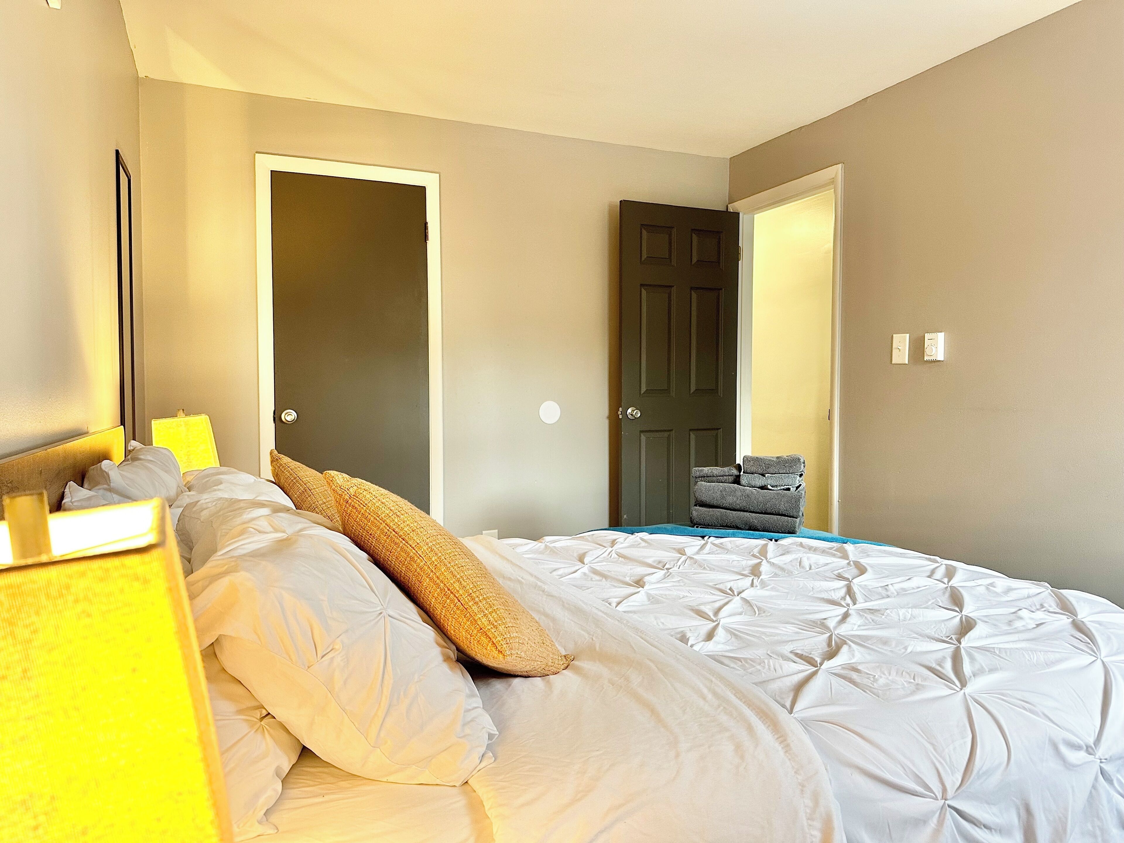 A neatly made bed with white linens and a few pillows, a bedside lamp, and a stack of folded towels on the floor.