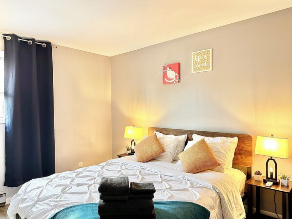 A hotel room with a bed, two bedside lamps, a nightstand with a lamp, a wall with two framed pictures, and a window with a curtain. - Comfy beds/Fast wifi/Getaway/Free Parking/Raging Waves Water park (Aurora)