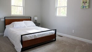 A bedroom with a wooden bed frame, white bedding, two windows, a nightstand with a lamp, and a wall-mounted painting.