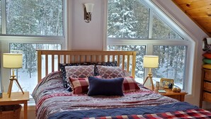 A bedroom with a wooden bed, two bedside lamps, a ceiling fan, and large windows with a snowy forest view.