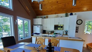 A wooden interior with a dining table set for four, a kitchen with white cabinets, and a large window providing natural light.