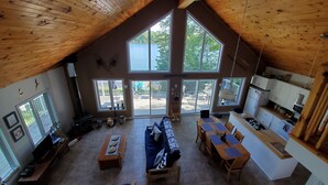 A spacious living area with a high ceiling, large windows, a fireplace, a dining table, and a kitchen area.