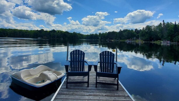 A wooden dock with two Adirondack chairs facing a calm lake, surrounded by trees and a clear sky.