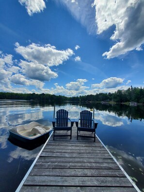 A wooden dock with two Adirondack chairs facing a calm lake, surrounded by trees and a clear sky.