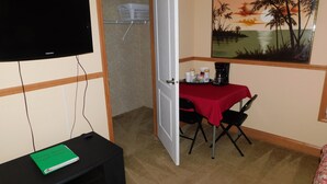A room with a TV, a desk with a red tablecloth, a chair, a bookshelf, and a painting on the wall.
