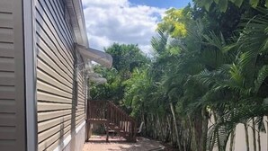 A brick pathway leading to a wooden deck, surrounded by lush greenery and a white wall.