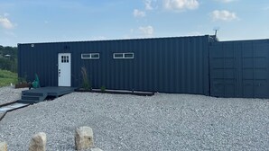 Een groot, donker gebouw in industriële stijl met een witte deur en ramen, gelegen op een grindpad met een kleine tuin ervoor.