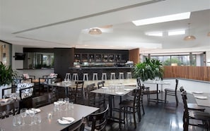 Restaurante moderno com pé-direito alto, área de bar e grandes janelas que proporcionam luz natural.