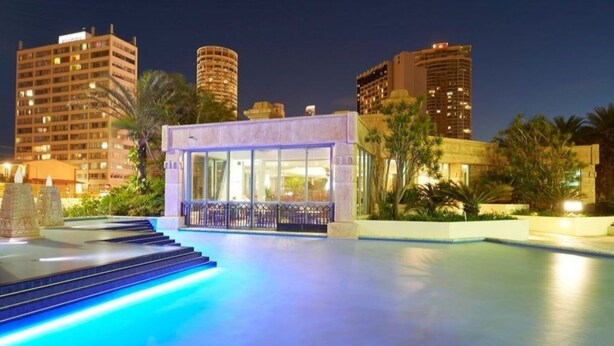 A modern hotel with a pool area, surrounded by palm trees and urban buildings at night. - Sun City - Q Stay (Surfers Paradise)