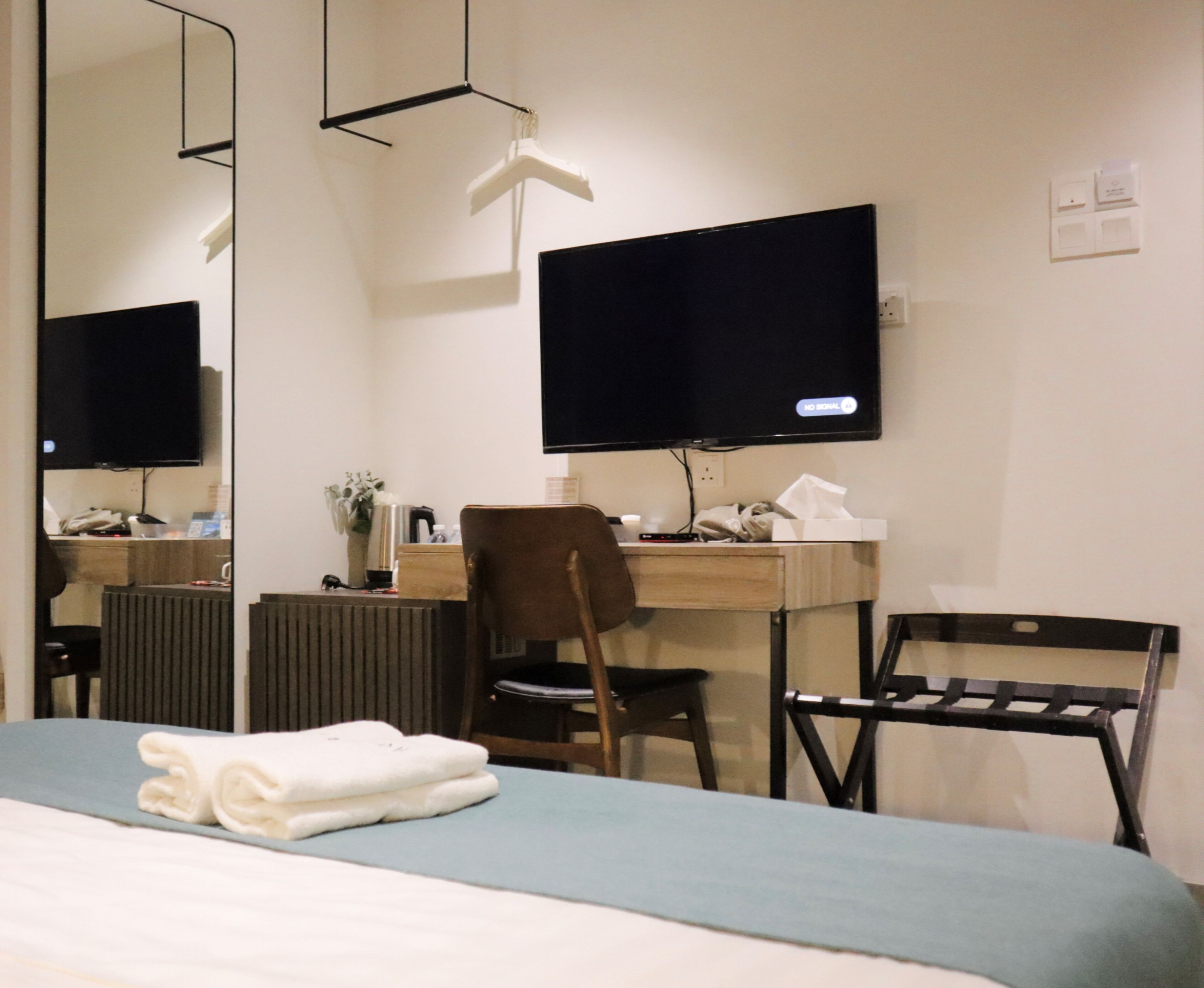 A hotel room with a bed, a desk with a computer, a chair, a radiator, and a TV mounted on the wall.