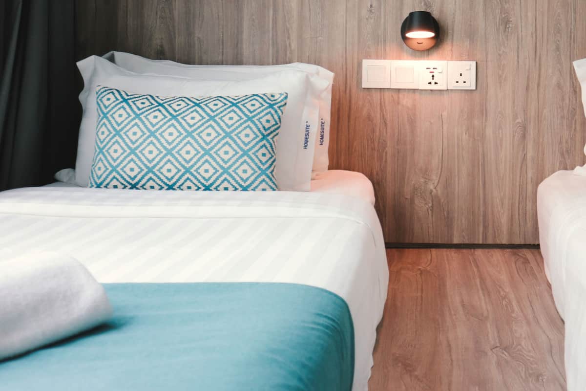Een hotelkamer met twee bedden, elk met een gekleurd kussen en een witte sprei. Er is een stopcontact en een wandlamp tussen de bedden.