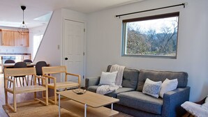 Een moderne woonkamer met een grijze bank, houten stoelen, een houten salontafel en een groot raam met uitzicht op bomen.