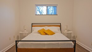 Een enkel bed met houten frame en witte beddengoed, twee nachtkastjes met lampen, een raam boven het bed en een effen witte muur.