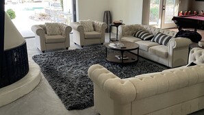 Ein geräumiges Wohnzimmer mit einem großen, beigefarbenen Sofa, zwei Sesseln, einem Couchtisch und einem Deckenventilator. Durch die Glastüren ist ein Pool zu sehen.