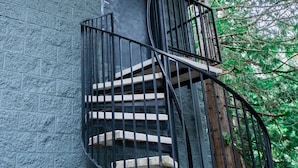 Un escalier en colimaçon avec des rampes noires menant à une porte en bois, adossé à un mur à texture et entouré de verdure.