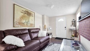 A living room with a brown leather sofa, a white fluffy pillow, a glass coffee table, a painting on the wall, and a brick fireplace. - Smart Saver Townhouse, Family Friendly, Close to NAS Jax, Long-Term, Washer and Dryer (Jacksonville)